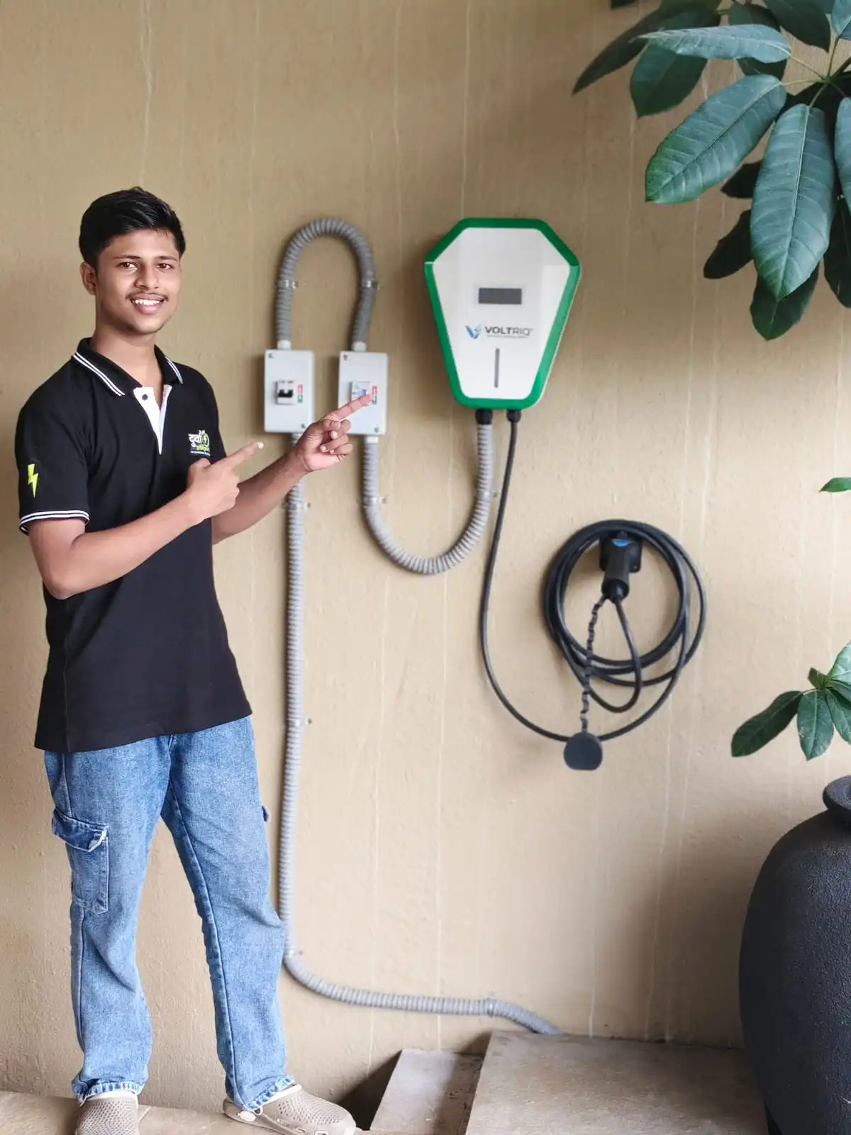 A Durva Electricals technician beside a completed Voltriq wallbox installation, pointing at the two protection enclosures mounted alongside it.