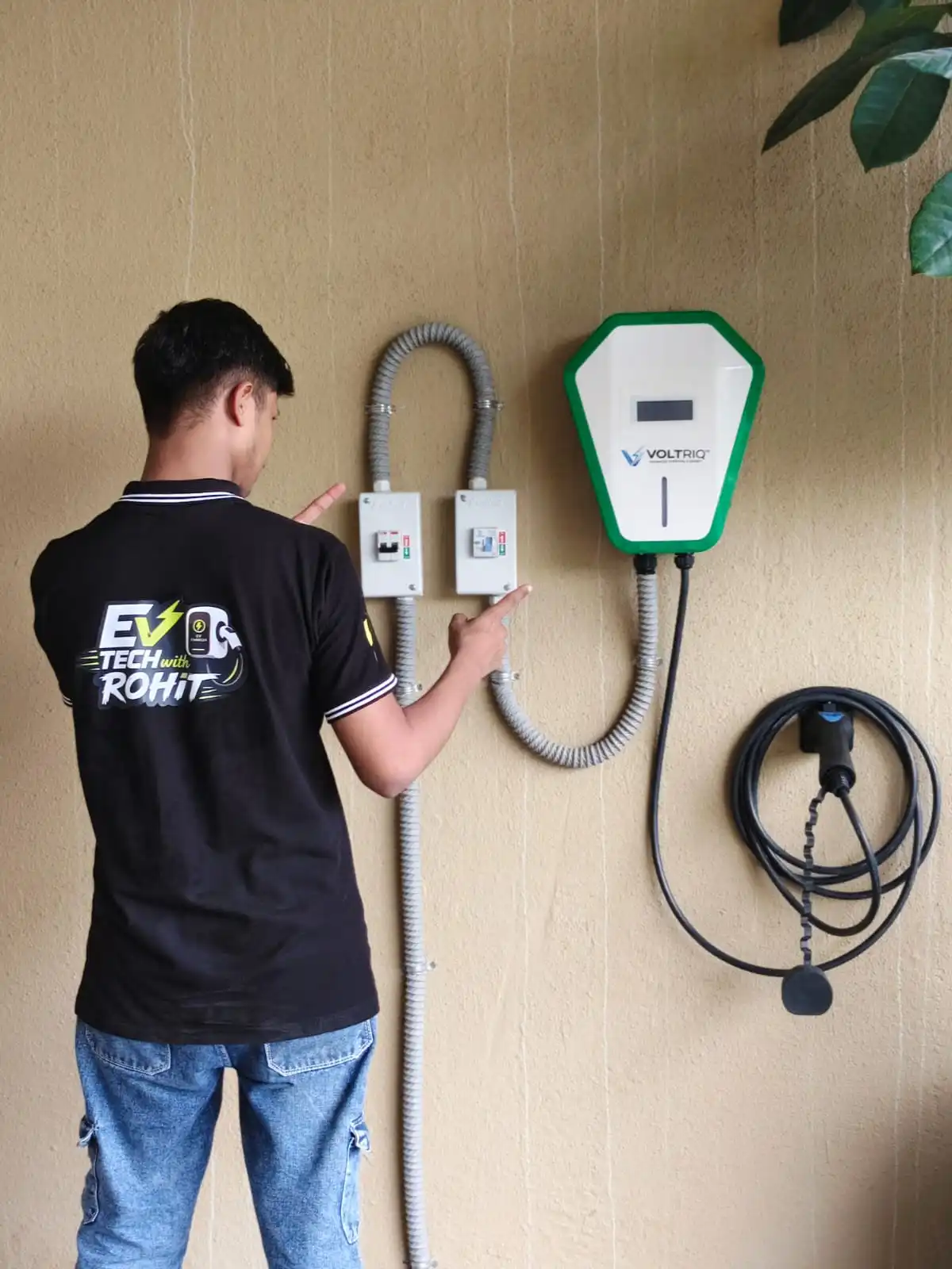 The same installation seen closer, with a technician pointing at the two separate protection enclosures beside the Voltriq wallbox and its Type 2 gun resting on a wall holster.