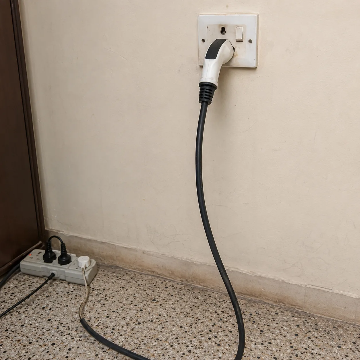 A portable EV charging cable plugged into an ordinary 15A wall socket, with an extension board on the floor beside it.